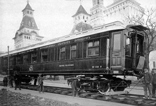 Historische Aufnahme eines luxuriösen Schlafwagens der CIWL mit Personen auf dem Bahnsteig, im Hintergrund Gebäude im russischen Stil.