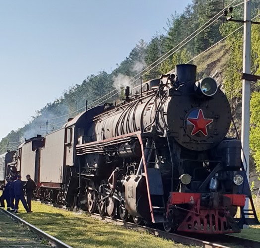 Dampflokomotive mit rotem Sowjetstern auf freier Strecke, im Vordergrund Eisenbahner, im Hintergrund bewaldeter Hang und Oberleitungen.