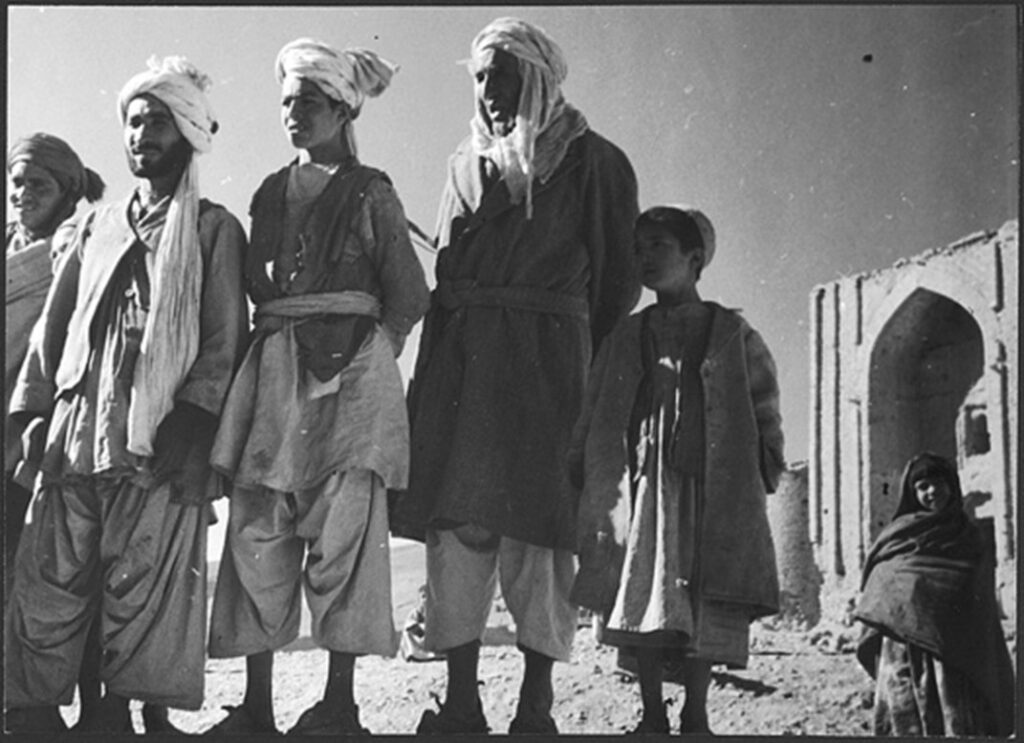 Gruppe von Männern und Kindern in traditioneller Kleidung steht vor einem alten, verfallenen Gebäude in einer Wüstenlandschaft.