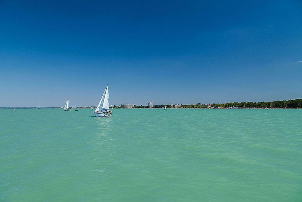 Segelboote auf dem türkisfarbenen Balaton bei klarem Himmel, im Hintergrund die Uferpromenade von Siófok.