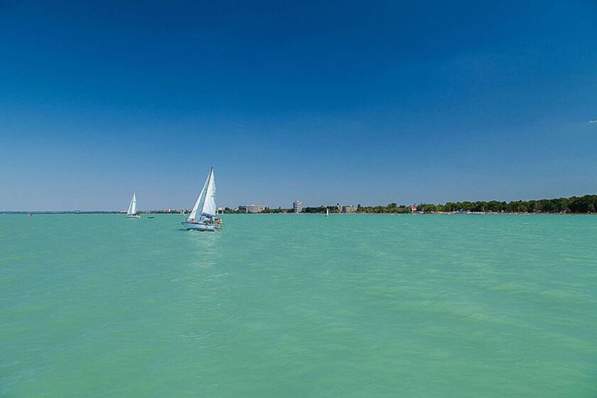 Segelboote auf dem türkisfarbenen Balaton bei klarem Himmel, im Hintergrund die Uferpromenade von Siófok.