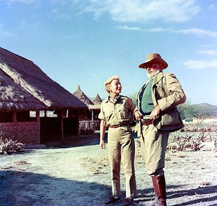 Mann und Frau in Safari-Kleidung stehen vor Strohhütten in trockener Landschaft unter blauem Himmel, scheinbar während einer Afrika-Reise.