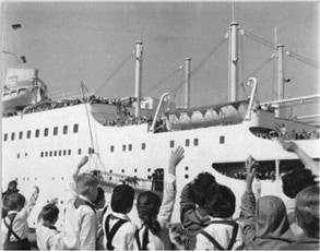Menschen winken einem ablegenden Passagierschiff; Blick auf das Schiff mit vielen Personen an Deck und Brücke im Hintergrund.