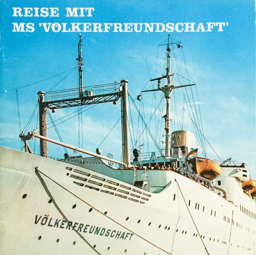 Großaufnahme des DDR-Passagierschiffs „Völkerfreundschaft“ unter blauem Himmel, mit sichtbarem Schriftzug am Bug und Menschen an Deck.