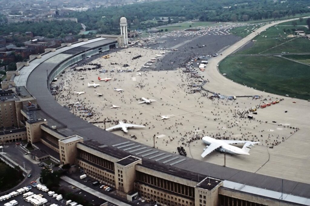 Luftaufnahme des Flughafens Berlin-Tempelhof mit Terminalbogen, Tower, abgestellten Flugzeugen und Menschenmassen auf dem Vorfeld.
