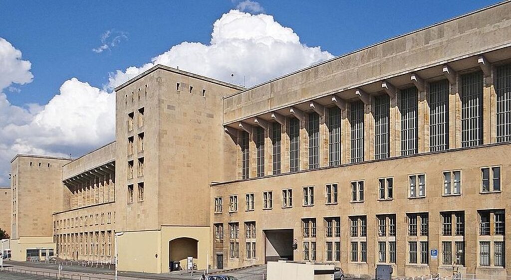 Moderne Außenansicht des Flughafens Berlin-Tempelhof: monumentale Sandsteinfassade mit hohen Fensterreihen vor blauem Himmel.