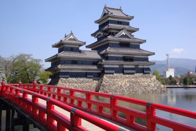 Japanische Burg am Wasser mit roter Brücke im Vordergrund unter klarem Himmel.