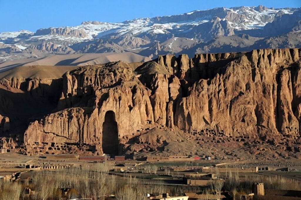 Felswand mit großer Nische der zerstörten Buddha-Statue von Bamiyan, davor Häuser und im Hintergrund schneebedeckte Berge.