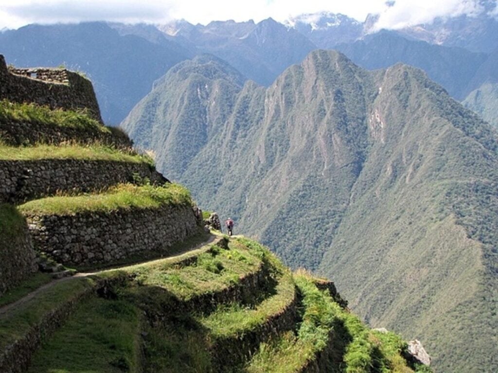 Steinterrassen an steilem Berghang mit schmalem Pfad vor hohen, bewaldeten Andengipfeln.