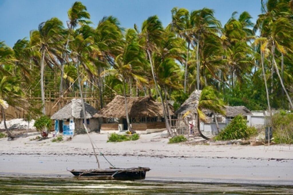 Palmengesäumter Sandstrand mit einfachen Hütten und kleinem Boot im flachen Wasser.
