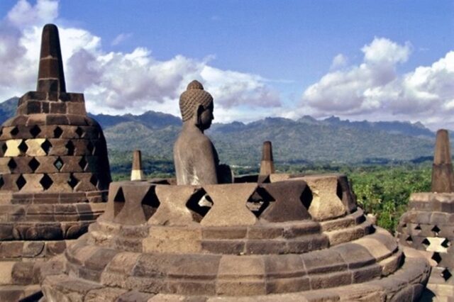 Buddha-Statue auf der Terrasse des Borobudur-Tempels vor grüner Landschaft, Bergen und wolkigem Himmel.
