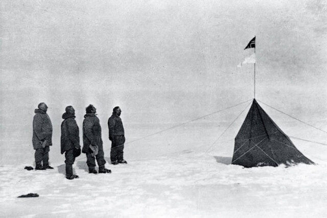 Vier Forscher stehen im Schnee vor einem Zelt mit Flagge in der Antarktis.