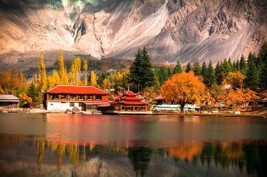Fotografie eines herbstlichen Sees mit traditionellem asiatischem Gebäude und bunten Bäumen im Vordergrund, umgeben von hohen Bergen im Hintergrund. Spiegelung von Gebäuden und Bäumen im ruhigen Wasser erzeugt harmonische und farbenfrohe Landschaftskomposition.