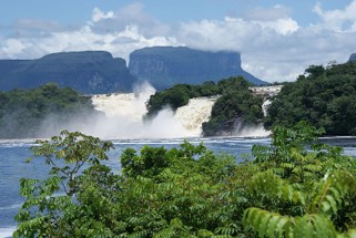 Fotografie einer großen Wasserfalllandschaft mit dichtem Grün im Vordergrund und Bergen im Hintergrund, die von Wolken teilweise bedeckt sind. Wasser stürzt kraftvoll über Felsen, erzeugt Gischt und vermittelt eine dynamische Naturszene.