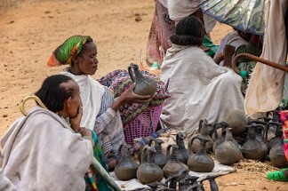 Fotografie zeigt mehrere Frauen in traditioneller Kleidung, die handgefertigte Tontöpfe auf einem Markt verkaufen. Töpfe sind in Reihen auf dem Boden angeordnet, Frauen sitzen oder kauern dahinter, Umgebung wirkt ländlich und trocken.