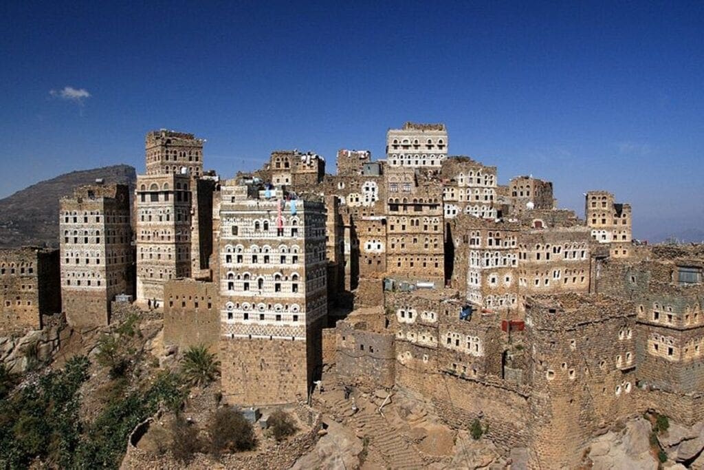 Fotografie einer traditionellen Bergsiedlung mit mehrstöckigen Lehmziegelhäusern und weißen Fensterrahmen, eingebettet in eine felsige Landschaft unter klarem blauem Himmel. Gebäude sind dicht aneinander gebaut, zeigen unterschiedliche Höhen und architektonische Details, die kulturelle Bauweise und Anpassung an das Gelände verdeutlichen.