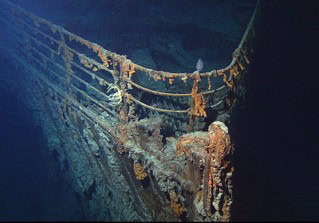 Fotografie eines versunkenen Schiffsbuges, der mit Korallen und Meeresbewuchs bedeckt ist. Aufnahme vermittelt Eindruck von Tiefe und Zustand des Wracks unter Wasser, mit sichtbaren Metallstrukturen und Seilen.